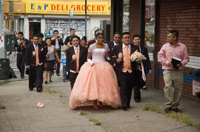 A bride and groom lead their wedding party down a block, with a mariachi quartet bringing up the rear.