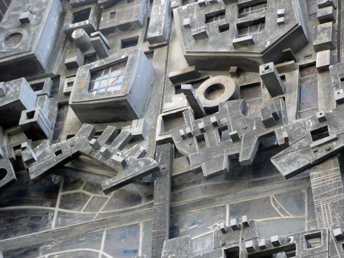 Building rooftops are seen from above, but they are islands in stained glass, a sculpture outside a church.