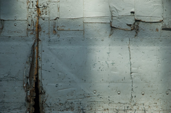 A weathered grey door shows craacks, peeling layers, and signs of several efforts to reinforce it.