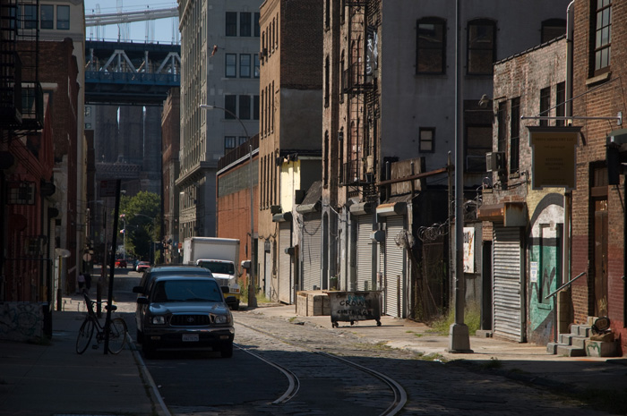 An old street has railroad tracks running down its center; storefronts are rolled down, and there are no signs of life. A bridge is seen at the far end.