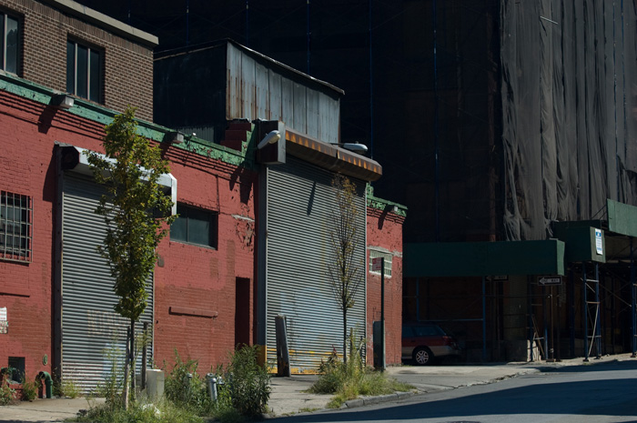 Harsh noon day sun hits rolled down garage fronts and casts deep shadows; in the background a building is covered in netting.