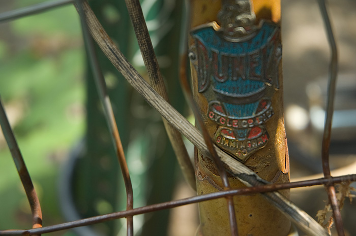 A worn, corroded bicycle head badge fights through basket wire and break cables for attention.