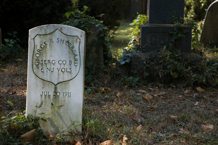 A simple white tombstone marks a veteran's grave in a cemetery with some overgrowth.