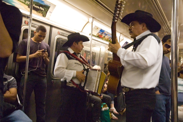 Two Mexican musicians, one with an accordion and the other with a guitar, play in the middle of a crowded NYC subway car.