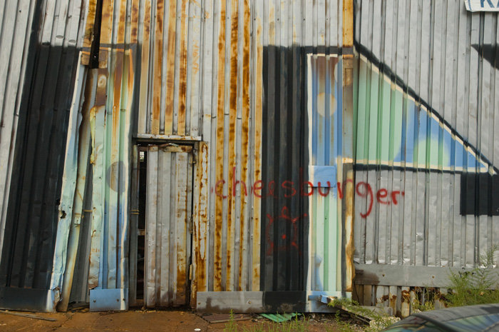 On a corrugated steel wall, already covered with grafffiti, the word 'cheeseburger' has been spray-painted in red.