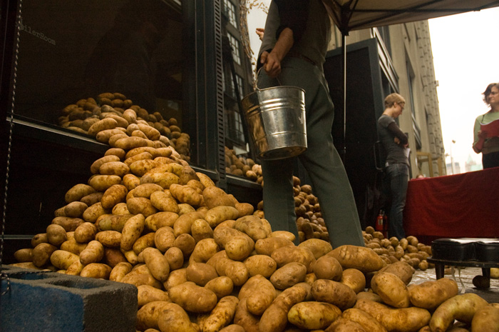 Piles of potatoes seem to pour through a storefront window, while a man stands nearby with a bucket.