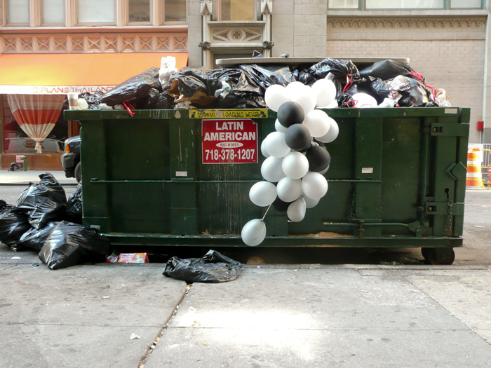 A dozen or so balloons, tied in a group, hang outside a dumspter.