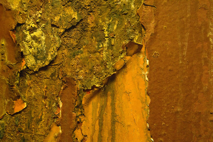Paint peels on a steel pillar on a subway platform, revealing layers of other colors as well as corrosion.