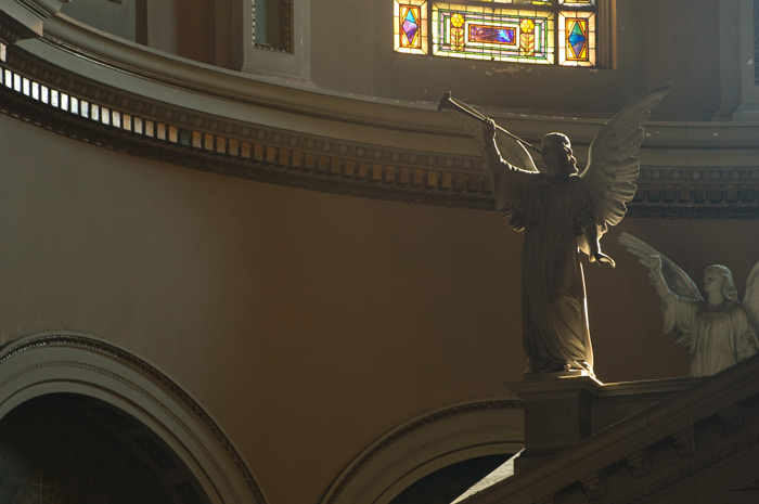 A statue of the archangel Gabriel, blowing his horn, is in near silhouette, atop a church altar.