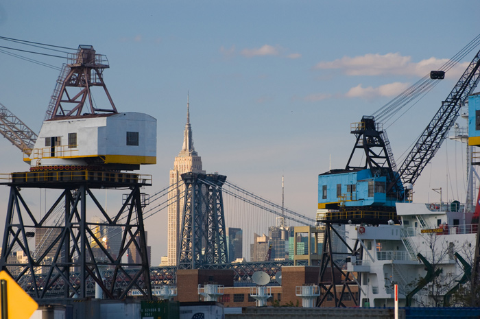 The tower of a bridge and the Empire State Building are framed by loading cranes.