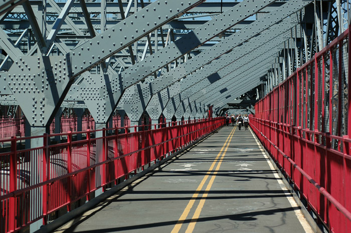 The asphalt of a bridge's pedestrian walkway is broken by sunlight and shadows from the reticulated structure.