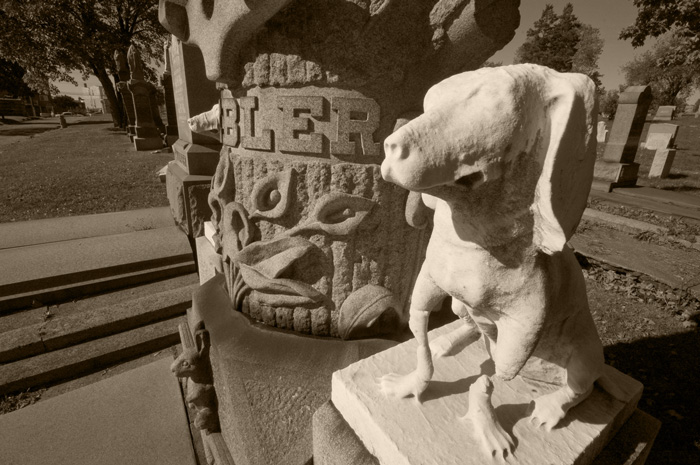 A statue of a hunting dog stands at attention next to a column over a tomb.