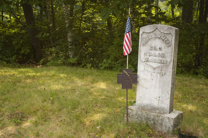 The tombstone of a Spanish American War veteran stands in a green graveyard, with a flag nearby.