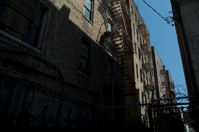 Shadows cover most of the back of an apartment building, but sunlight creeps through to light fire escapes and barbed wire.