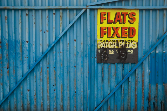 A blue corrugated metal fence has a hand-painted yellow sign with red letters, advertising tire repair.