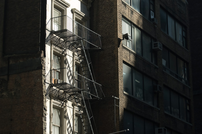 A fire escape is highlit by a beam of sunlight; the rest of the street is in shadows.