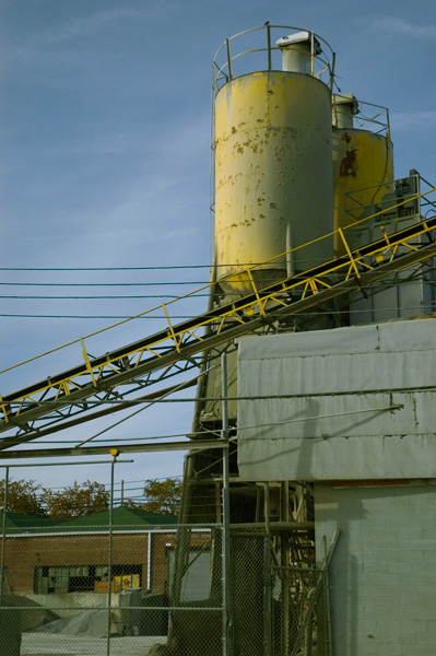 The yellow, towering silos and conveyor belt of a cement factory stand out against a blue sky.