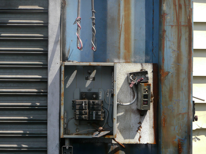 Sunlight and shadows mark an open circuit breaker box; cables hang down.