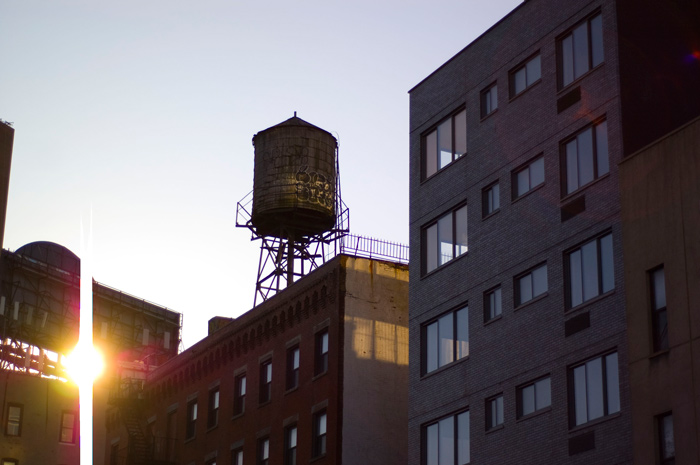 A ray of sunshine squeezes through billboards, and in the brightness a rooftop water tank stands in silhouette.