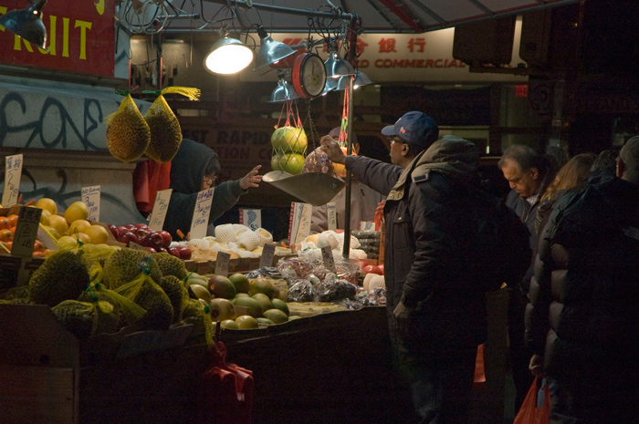 A light shines on a fruit stand at night, where a customer lifts his fruit to scale and a cashier tallies purchases.