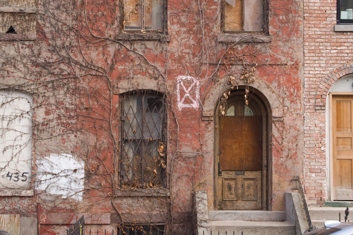 A brick home is about to undergo rehab, but dead ivy covers its front, and a spray-painted symbol warns of structural instability.