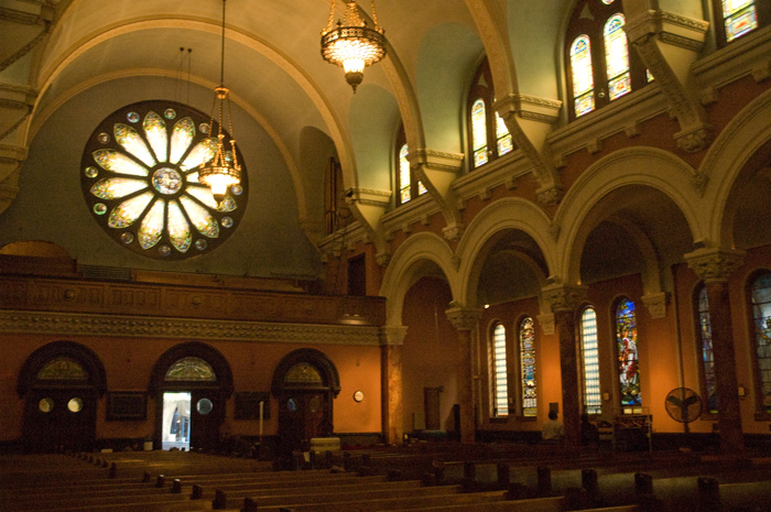 A church interior has two tiers of stained glass windows, and a large rose window at the rear.