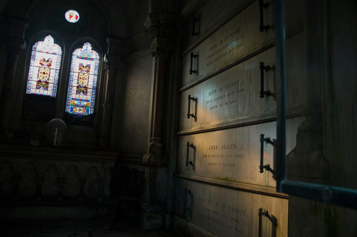 Light from stained glass windows shines on tombs in an otherwise dark mausoleum.