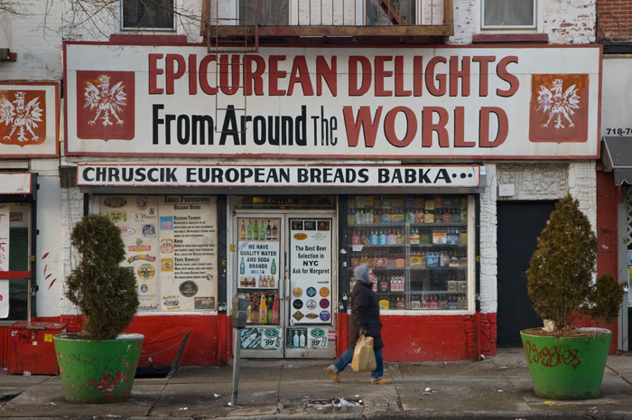 A woman with a shopping bag walks past a Polish grocer, whose storefront is painted in Red, White, and bold black letters and decorated with Polish eagles.