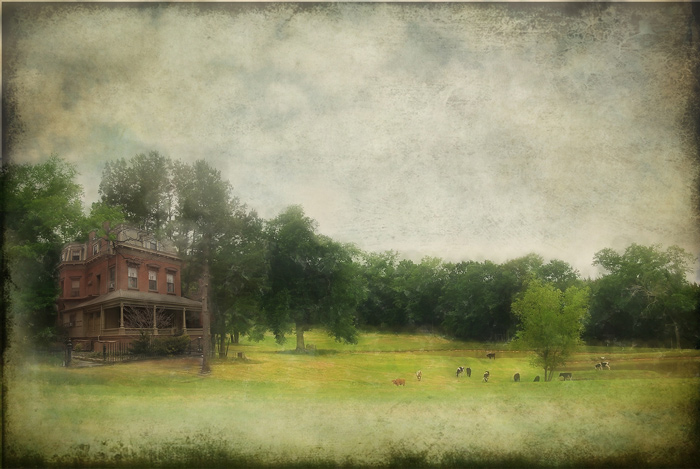 A brown, multi-story house with a porch sits on a farm, with green fields and scattered livestock.