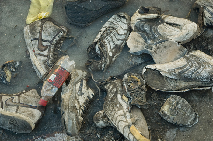 A pile of dirty, worn, torn sneakers sits in a frozen puddle, with other garbage.
