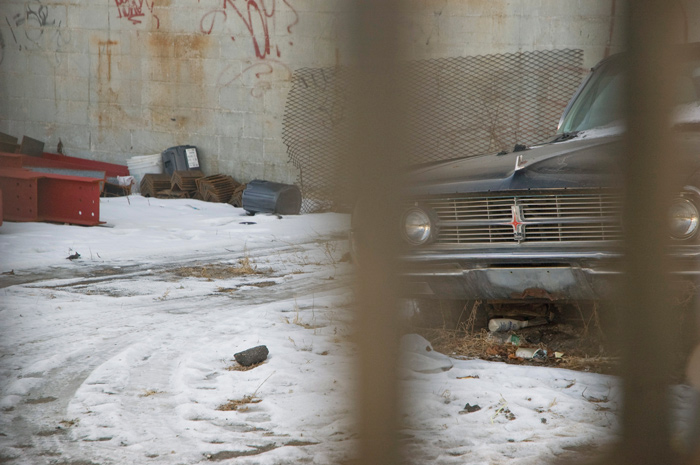 An old car, with its grille and medallion in clear focus, peeks between bars of a fence; junk litters the yard.