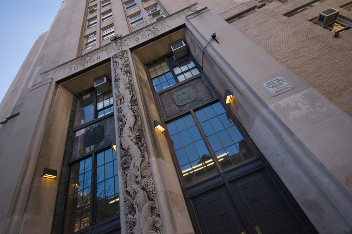 The photo shows a sweeping view of a building up to the sky; the building front is engraved with trees, vines, and the school name, named for Joan of Arc.