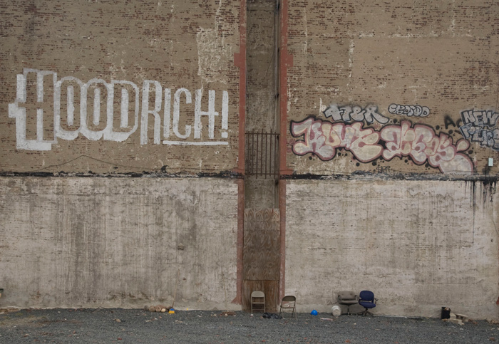 A brown, blank wall bears graffiti where a missing building's rooftop was. There are old chairs and other debris.