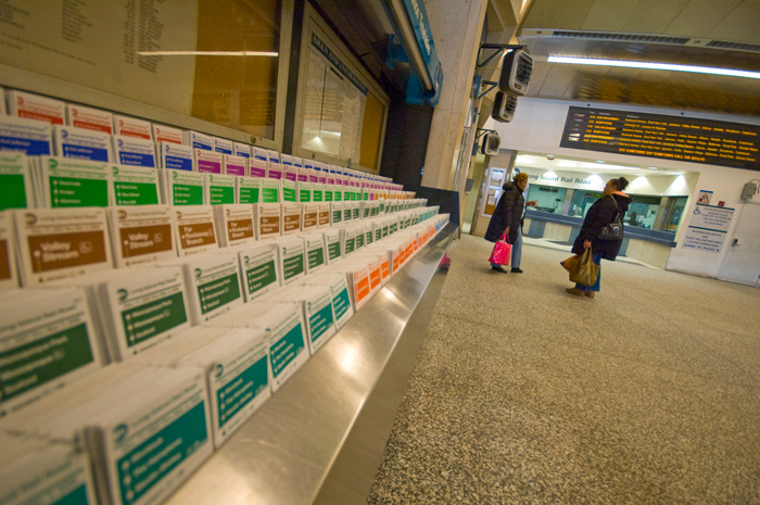 Racks of railroad timetables lead back to two travelers and ticket windows.