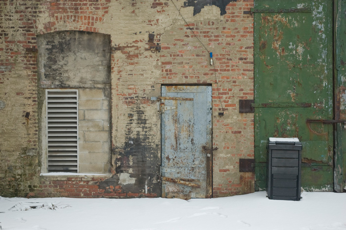 A metal door and other grates are in an old brock wall, and snow is on the ground.