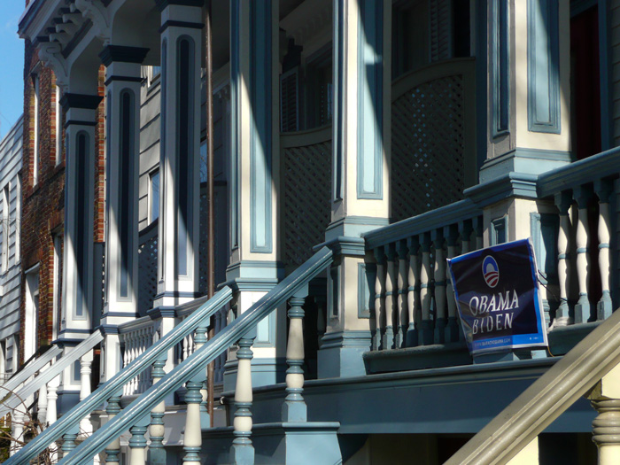 A banner for Barack Obama and Joe Biden hangs on the wooden railings of a porched house, painted blue and white like others nearby.