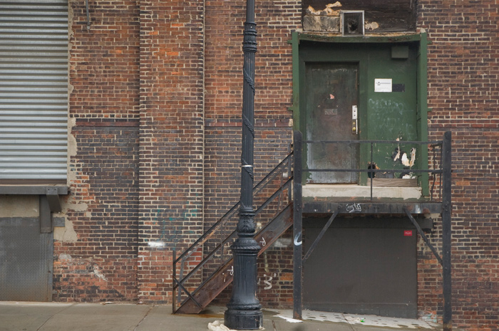 A black metal staircase on the side of an old brick building leads to a green door.