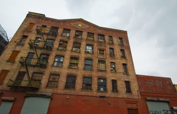 The windows of an old brick industrial building have been boarded up or broken, but an ornamental diamond remains at the top.