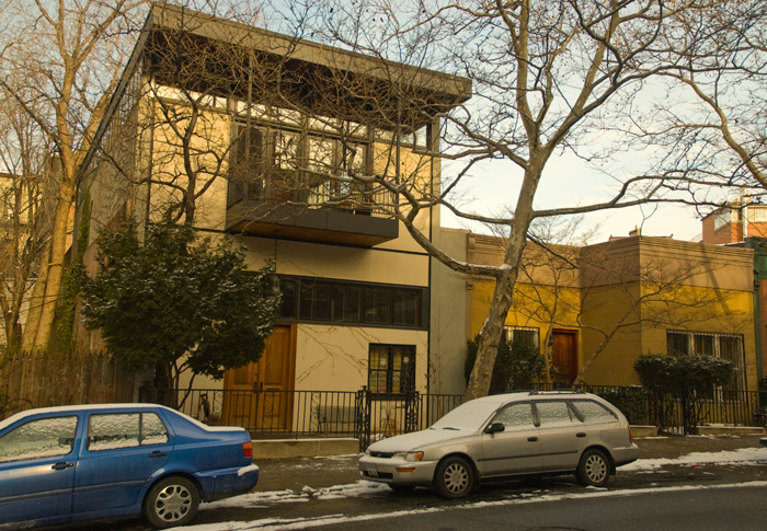 A building shaped like a block, with clean lines and no ornamentation, has a metal balcony and a jutting flat eave.