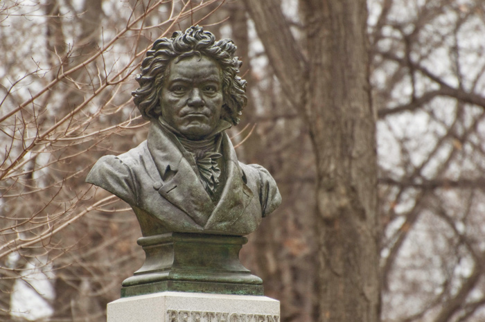 A bust of Beethoven sits on a pedesatl, in front of trees which have lost all their leaves.