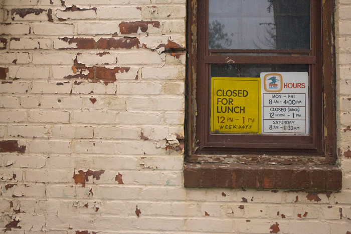 Two signs in a window announce the post office's hours, including lunch hour closing. The paint on the wall is pealing.