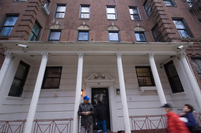 A man stands by the pillars of his building's porch, watching passersby.