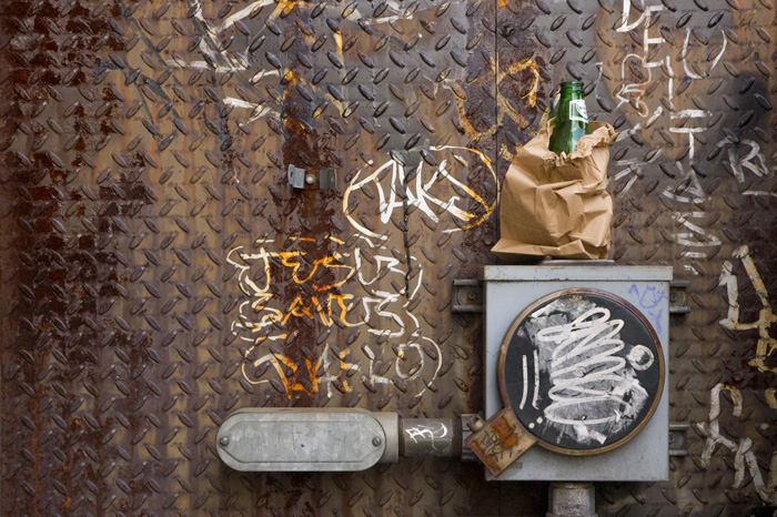 A beer bottle rests on top of an electrical box covered in graffiti, with 'Jesus Saves' scrawled nearby.