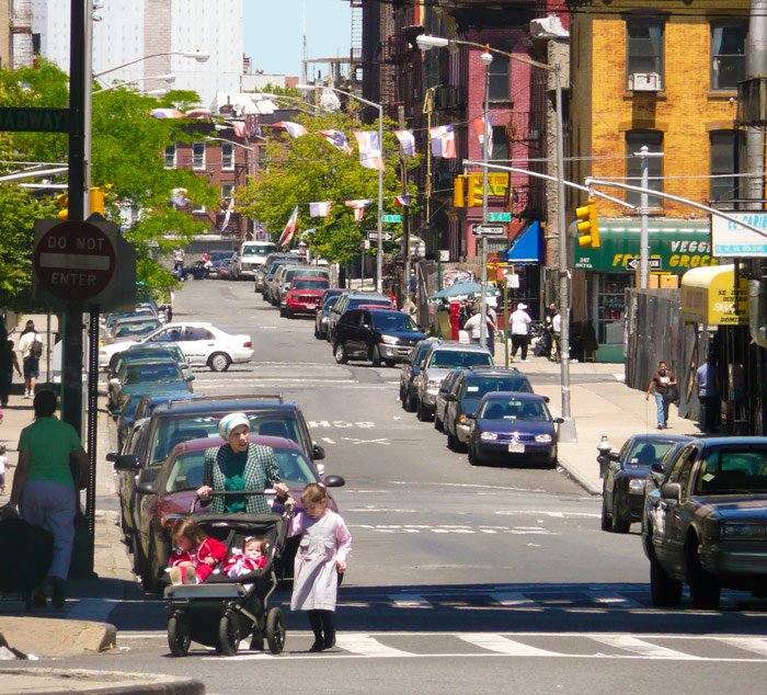 A mother pushes a stroller with two children on a city street, while a third walks alongside.