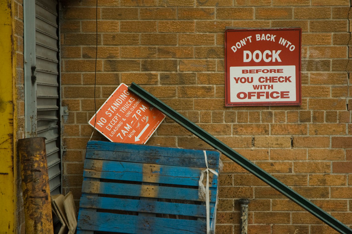 A sign warns truck drivers to check with the office before backing into the loading dock; next to it, another sign has been toppled.