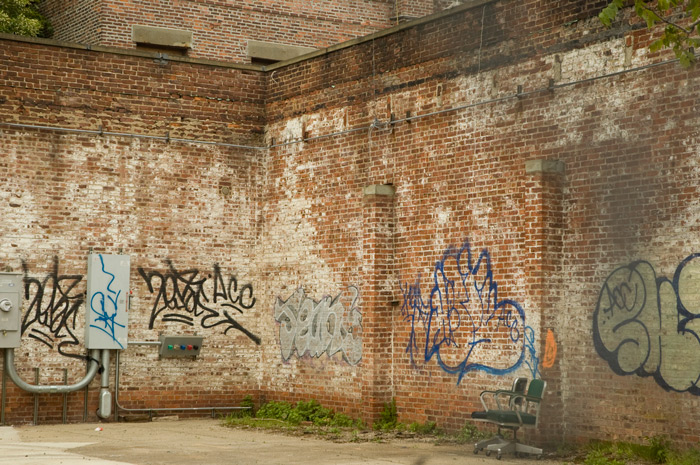 Two graffiti-covered brick walls join in courtard, where two office chairs sit.