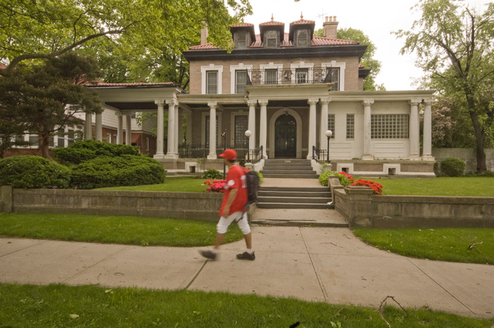 A baseball player walks in front of a mansion with columns, multiple stories, and bay windows, on a tailored green lawn.