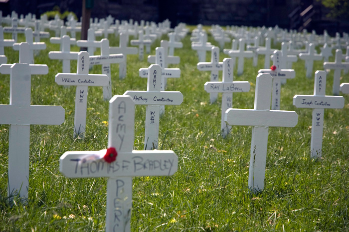 Small white, wooden crosses, hand-painted with the names of fallen soldiers, fill a green church lawn.