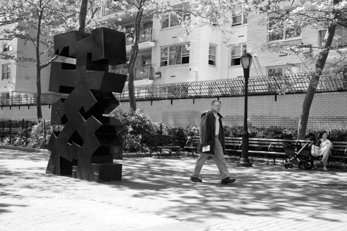 A man walks past a tall, black abstract sculpture in a tree-lined plaza.