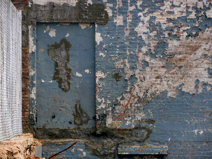 The side of a building has a walled up door, lots of peeling paint, and crumbling concrete.